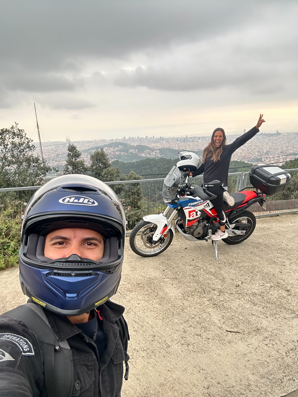 Passenger standing on the footpegs with her arm raised at a lookout above Barcelona