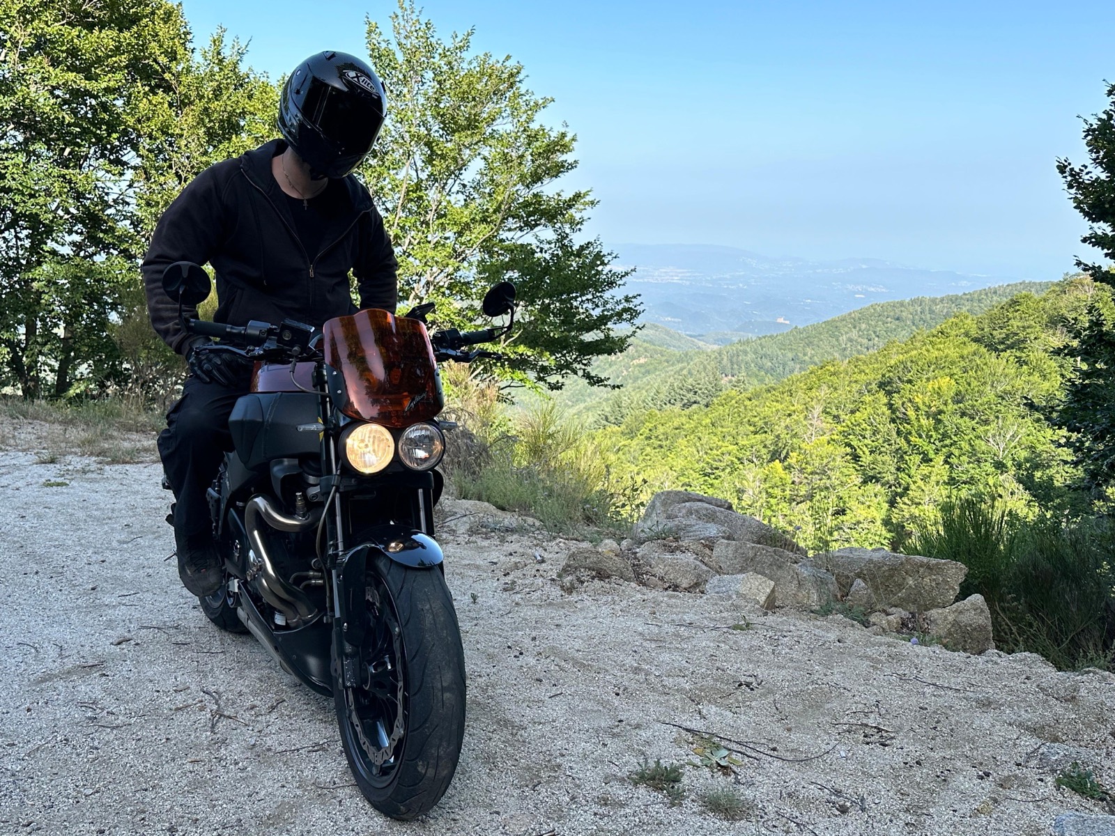 A rider on a black motorcycle at a viewpoint in the Montseny, forest below and the sea on the horizon