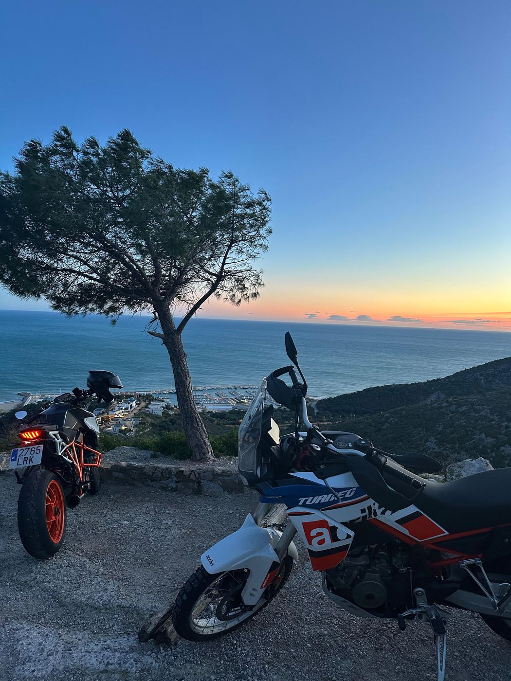 Two motorcycles parked at a viewpoint on the Garraf, a pine tree and the sea at sunset behind them